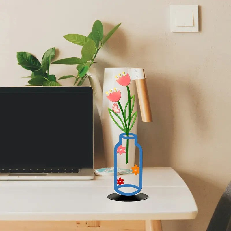 Colorful wall vase decoration with flowers on a desk beside a laptop and green plants.